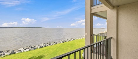 A balcony with a view of a calm body of water, a grassy area, and a distant shoreline.