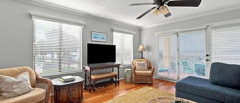 A living room with a ceiling fan, a television, wicker furniture, and large windows with blinds.