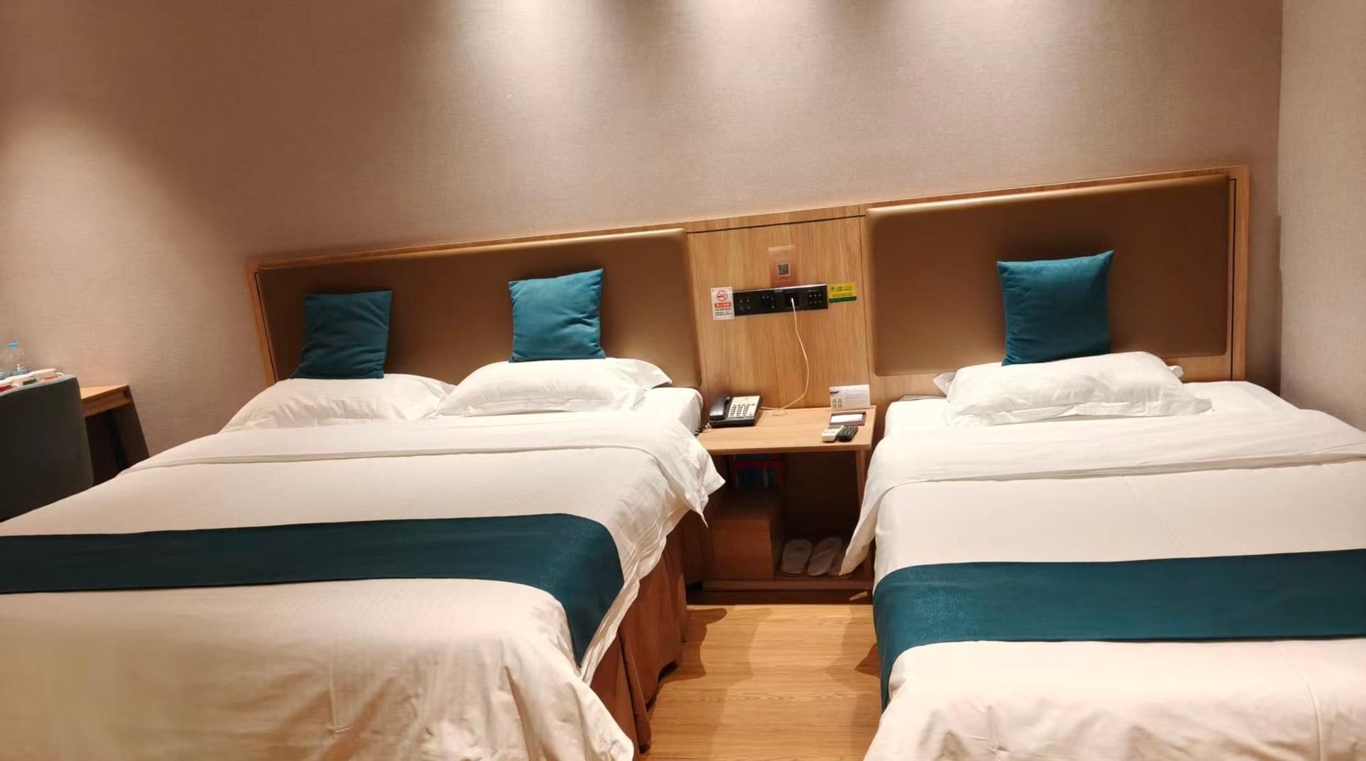 A hotel room with two beds, each with white bedding and teal pillows. There is a wooden headboard, a bedside table with a phone, and a wall-mounted control panel.