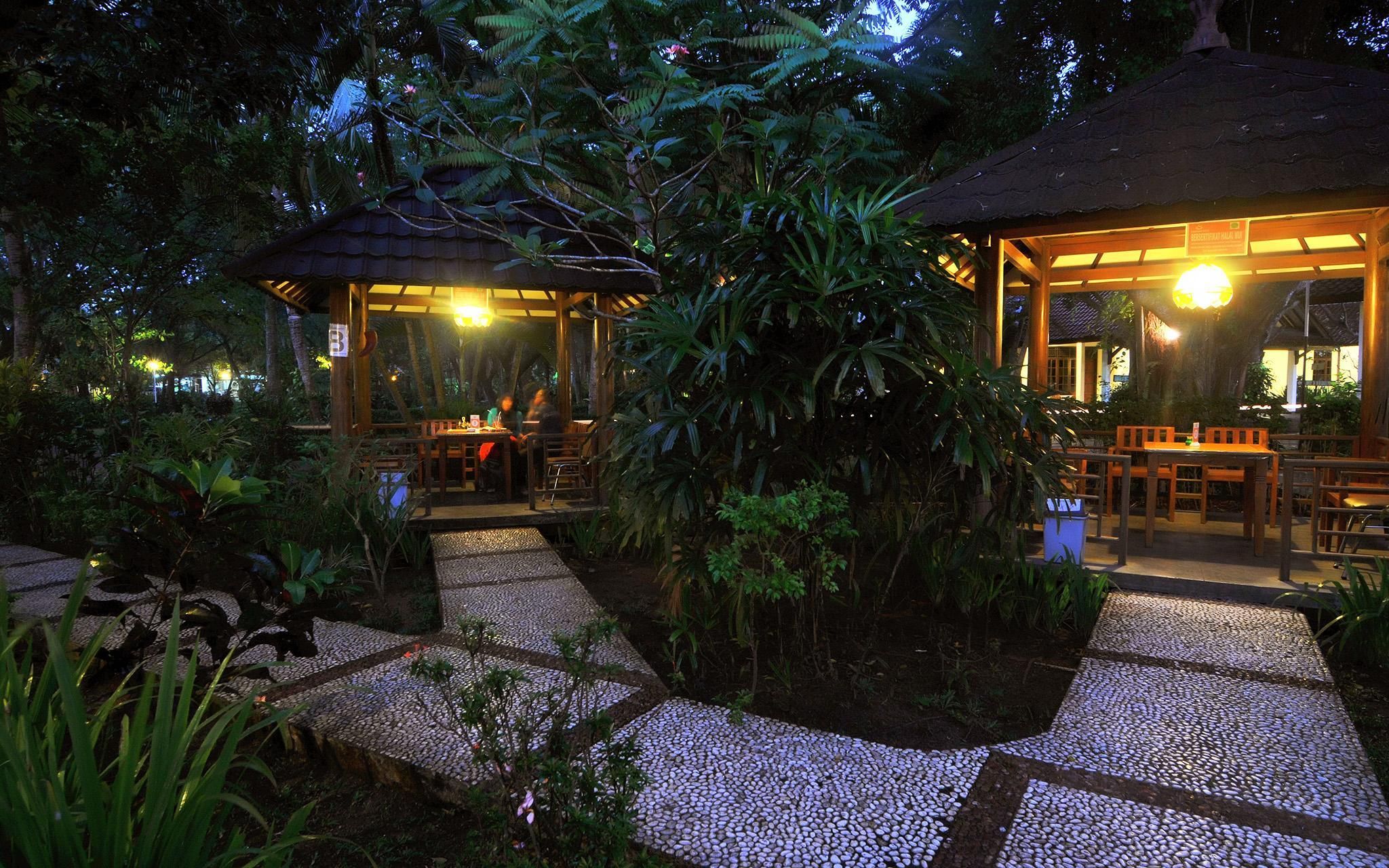 An outdoor dining area with tables and chairs, surrounded by lush greenery and a thatched-roof structure.