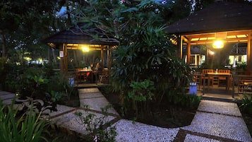 An outdoor dining area with tables and chairs, surrounded by lush greenery and a thatched-roof structure.