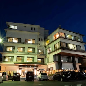 Un hotel de varios pisos con diseño moderno, que cuenta con balcones en los pisos superiores y un estacionamiento al frente.