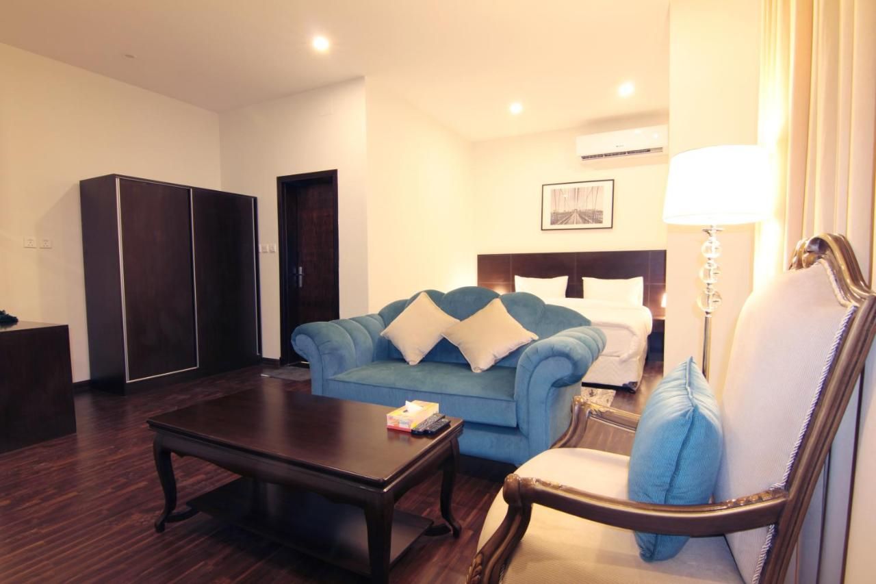 Una habitación de hotel con un sofá azul, una mesa de centro de madera, un sillón blanco, una cama con ropa de cama blanca y una mesita de noche con una lámpara.