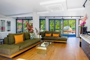 A modern living room with a sectional sofa, a glass coffee table, and large windows offering a view of greenery and buildings. - Nobile Boutique Hotel (Corfu)