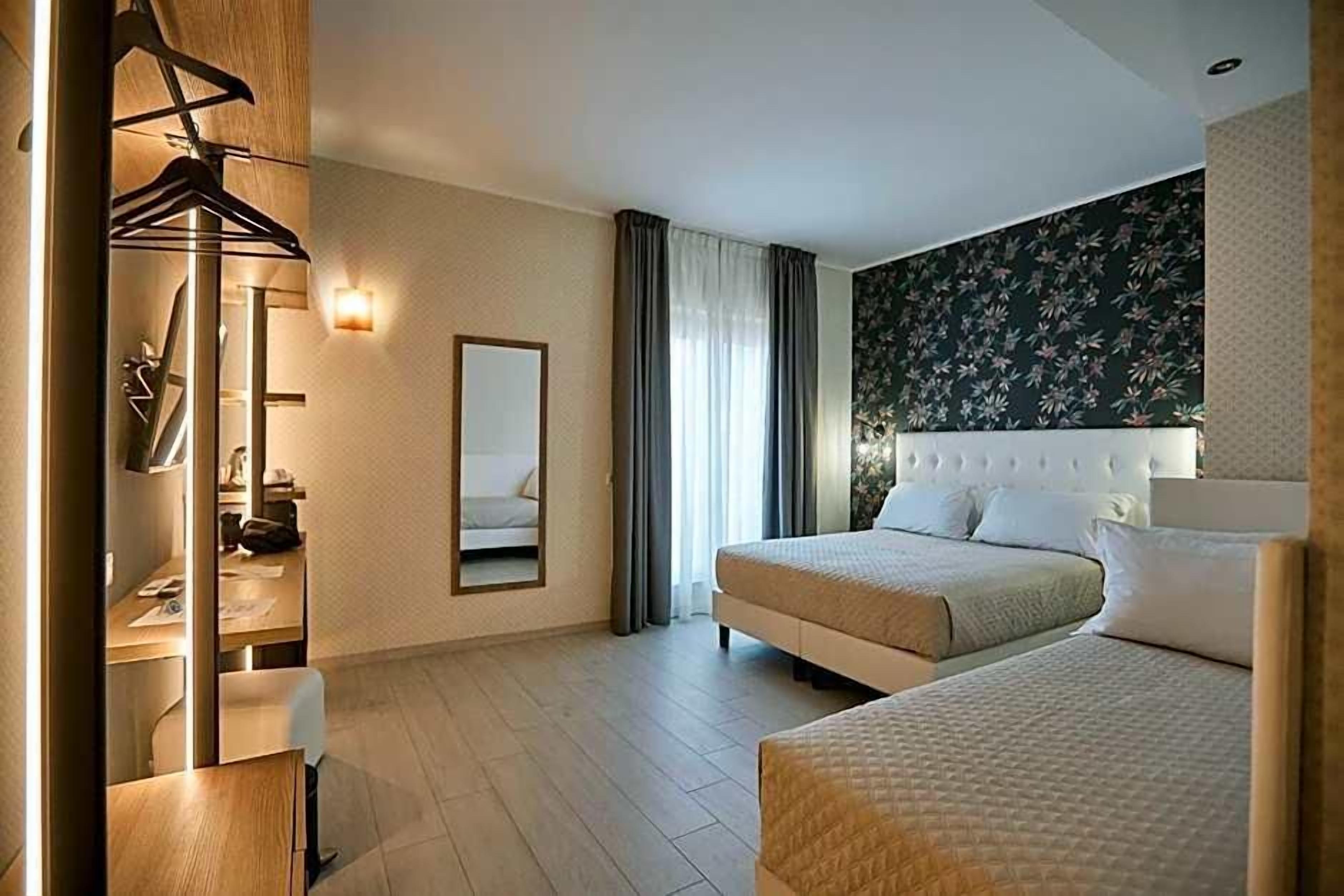 A hotel room with two beds, a wardrobe, a mirror, and a wall with a floral pattern.