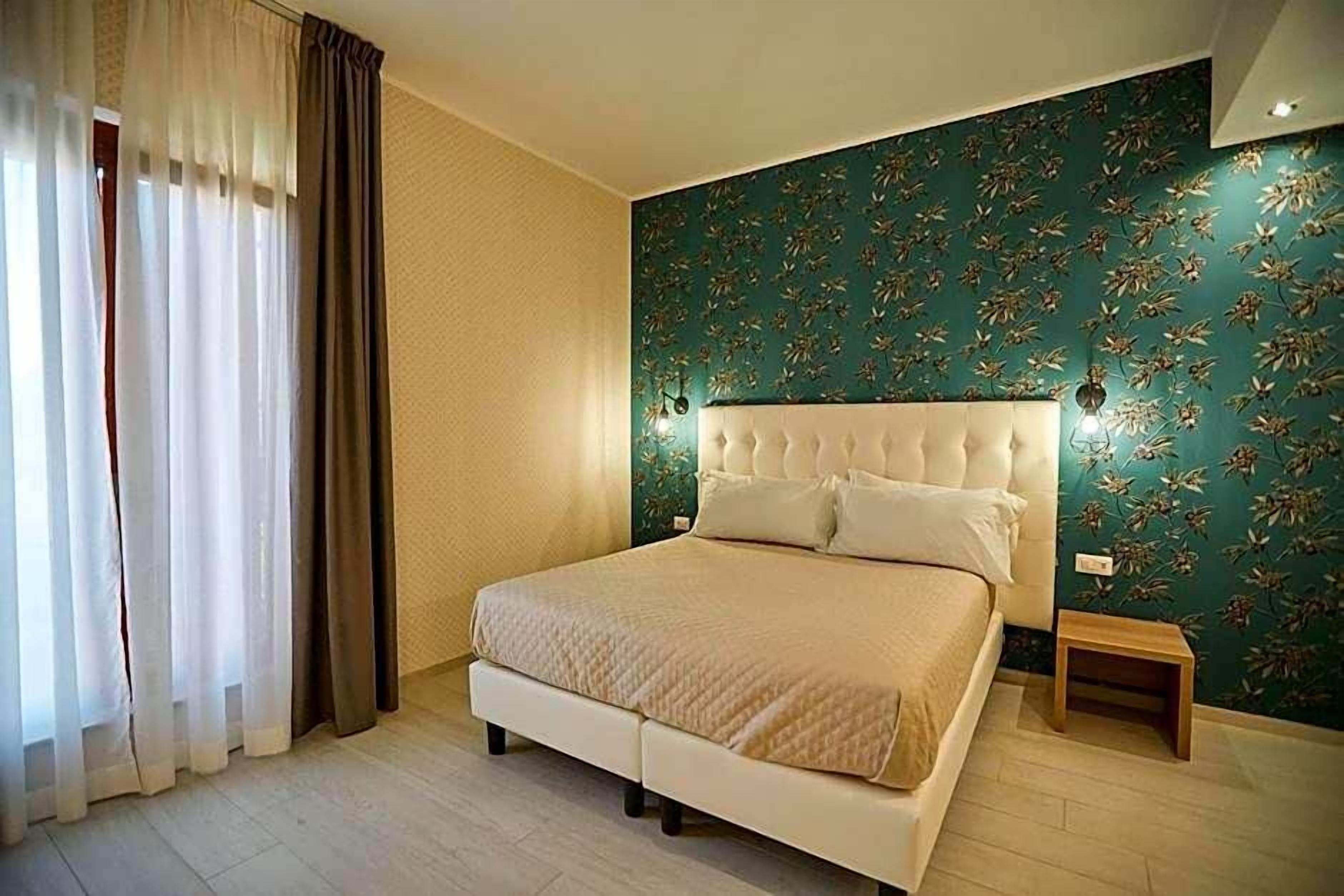 A bedroom with a bed, curtains, a nightstand, and a wall with a floral pattern.