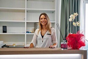 A reception desk with a woman standing behind it, a shelf with books and a clock, and a vase with flowers and a candle. - Castrichella Boutique Hotel (Fiumicino)