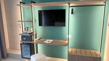 Quarto compacto com cama, pequena escrivaninha, televisão e mini-geladeira.