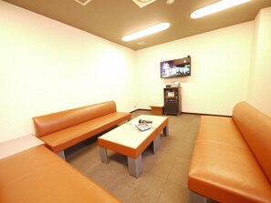 A waiting area with a television mounted on the wall, a vending machine, and two orange sofas. - Route-inn Grantia Wakamiya (Miyawaka)