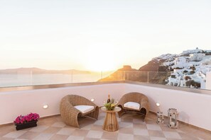 Ein Balkon mit Korbstühlen, einem kleinen Tisch mit Blumen und Blick auf weiße Häuser im Abendrot.