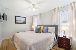 A bedroom with a bed, a ceiling fan, a TV, a window with curtains, and a bedside table with a lamp. - Gallery Stays - Low Country (Savannah)