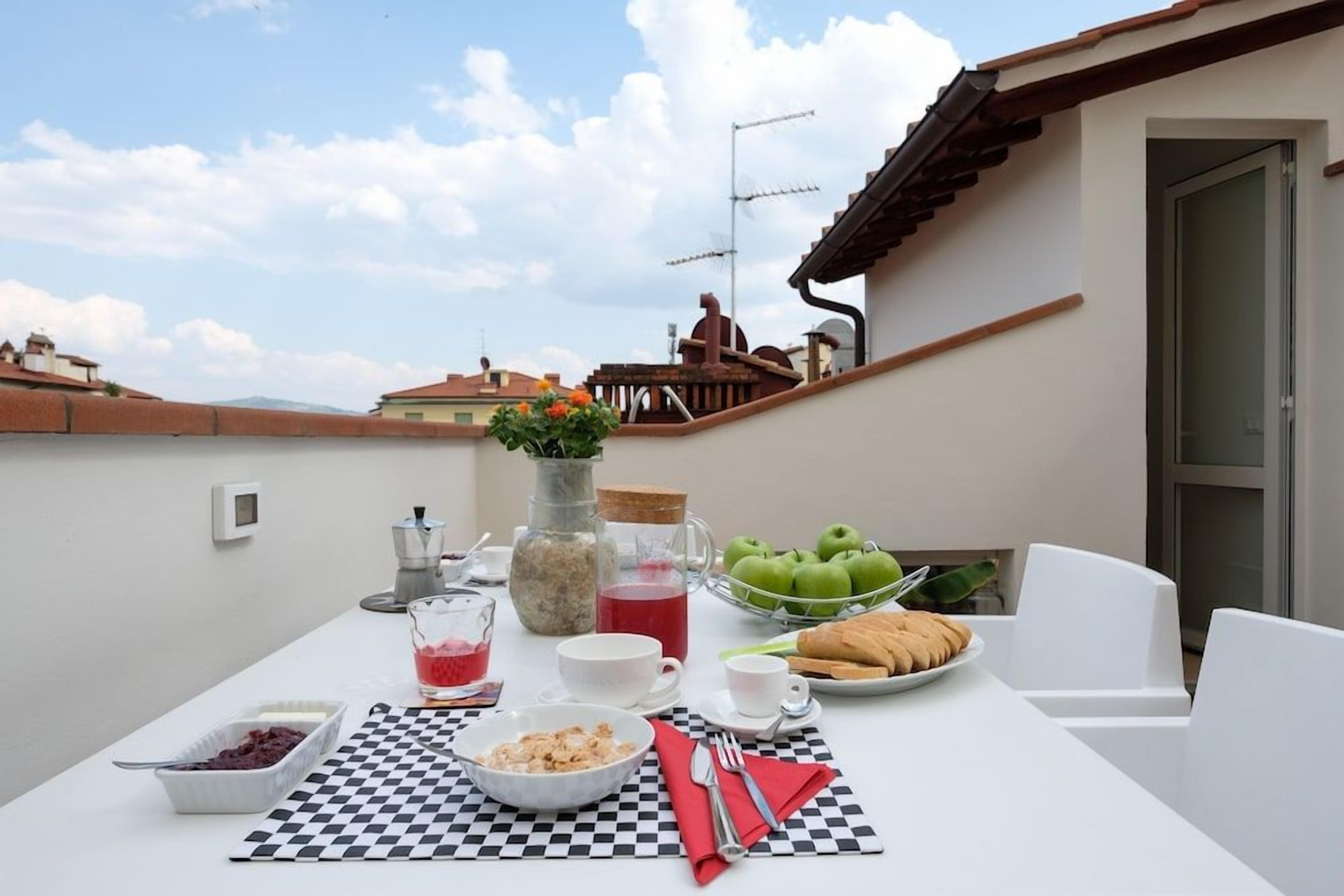 Una mesa servida con alimentos para el desayuno, como frutas, pan y bebidas, con vista al cielo y edificios al fondo.