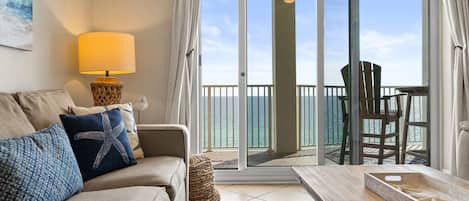 A living room with a sofa, a lamp, a wooden table, and a view of the ocean through a sliding glass door.