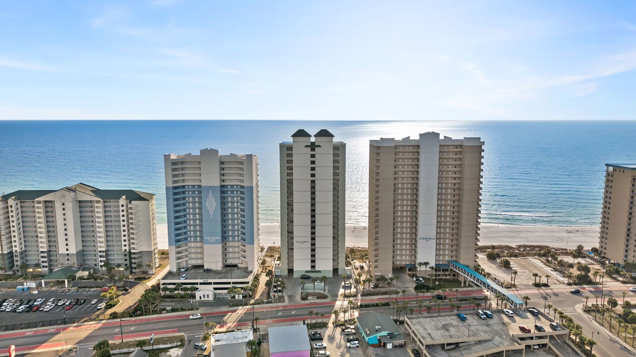A beachfront hotel complex with multiple high-rise buildings, a parking area, and a road leading to the beach.
