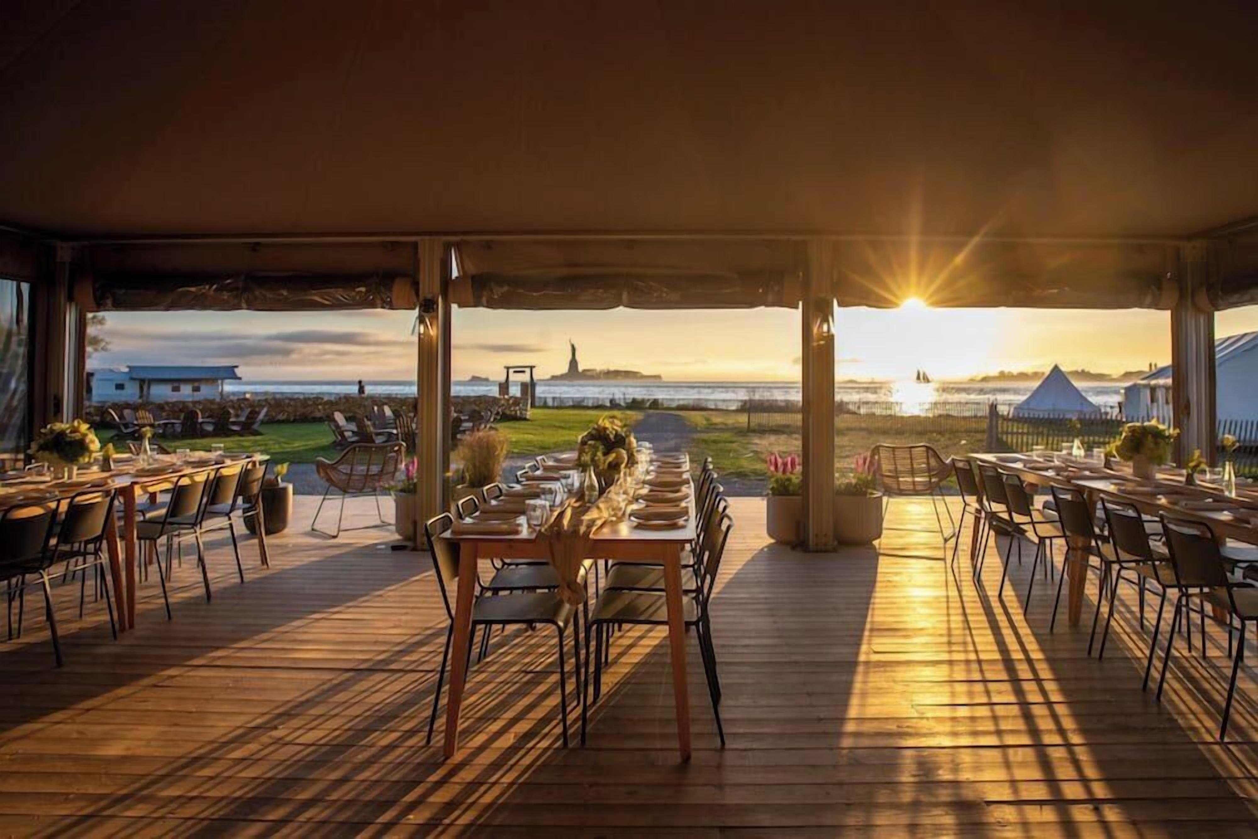 An outdoor dining area with tables and chairs, a view of the Statue of Liberty, and a sunset.
