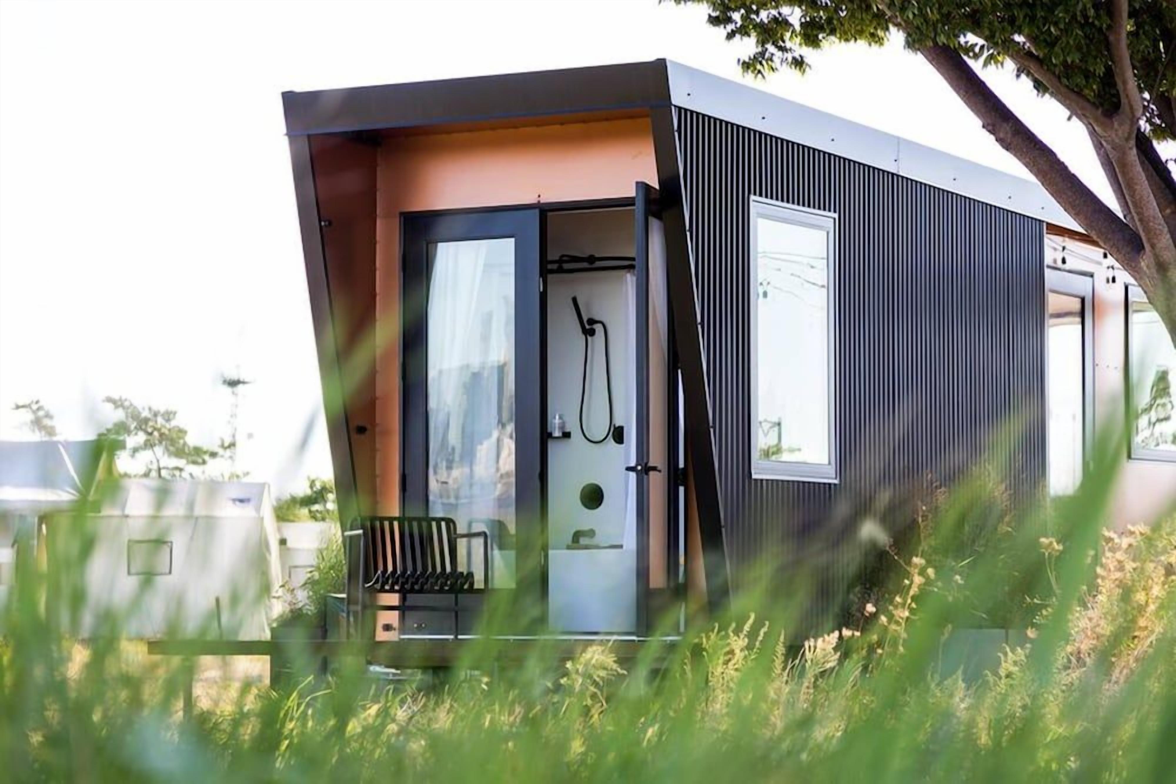 A modern, compact mobile home with a sliding door entrance, a small porch with a bench, and a window overlooking a grassy area.