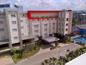 Un hotel de varios pisos con un detalle rojo en el nivel superior, piscina y un jardín ajardinado.