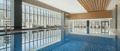 A large indoor swimming pool with clear glass walls, a modern ceiling design, and multiple lounge chairs along the poolside.