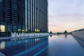 Um edifício moderno com fachada de vidro refletante, piscina infinita no terraço com espreguiçadeiras e céu claro.