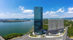 A modern high-rise building with a glass facade, adjacent to a body of water and a bridge, surrounded by greenery and other buildings. - Crowne Plaza Chaozhou Riverside (Chaozhou)