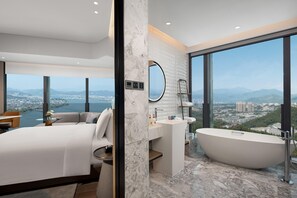 Quarto de hotel moderno com uma cama grande, banheiro com banheira independente e vista para a cidade através de grandes janelas.