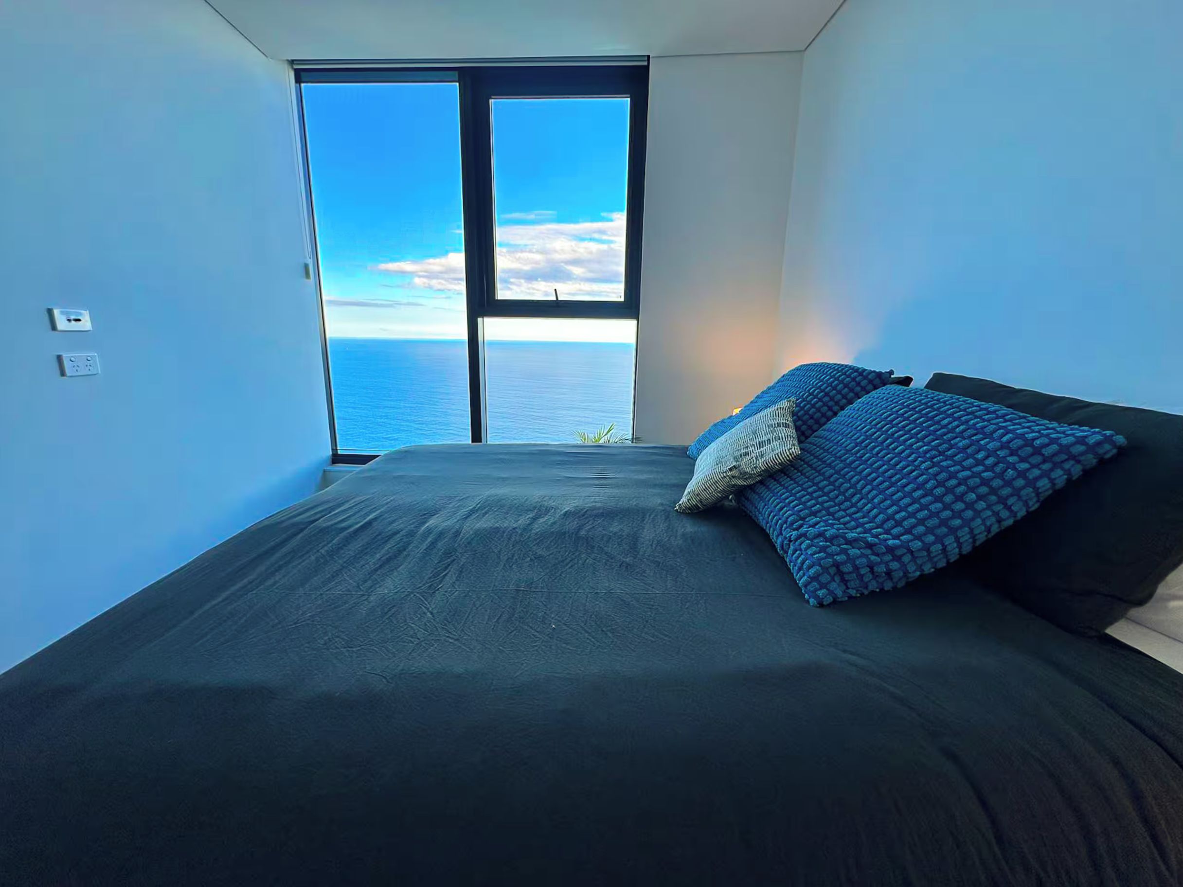 A bedroom with a large bed, a view of the sea through a window, and a power outlet on the wall.