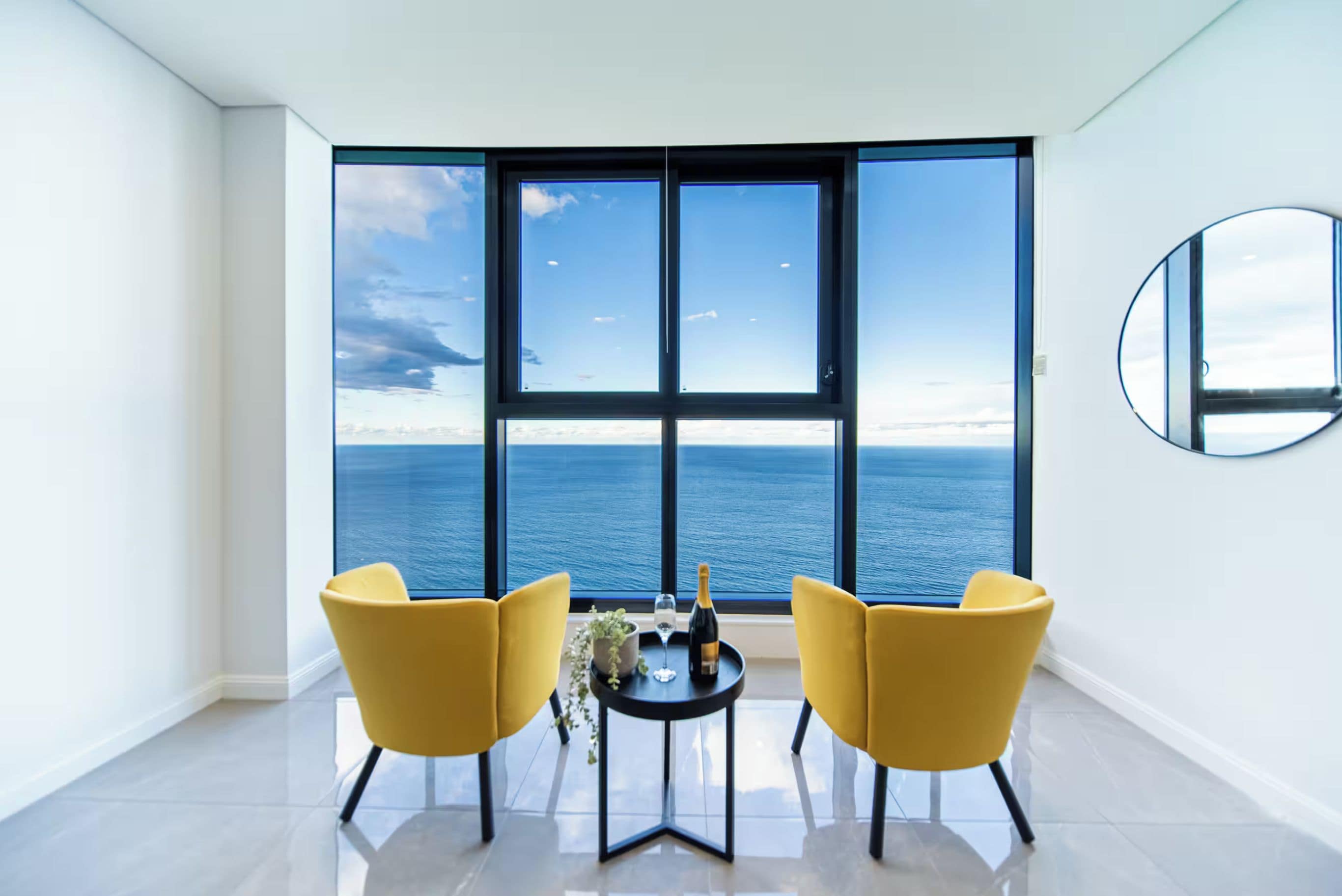 Une chambre moderne avec une grande fenêtre donnant sur la mer, deux chaises jaunes, une petite table avec une bouteille et un verre, et un miroir rond accroché au mur.