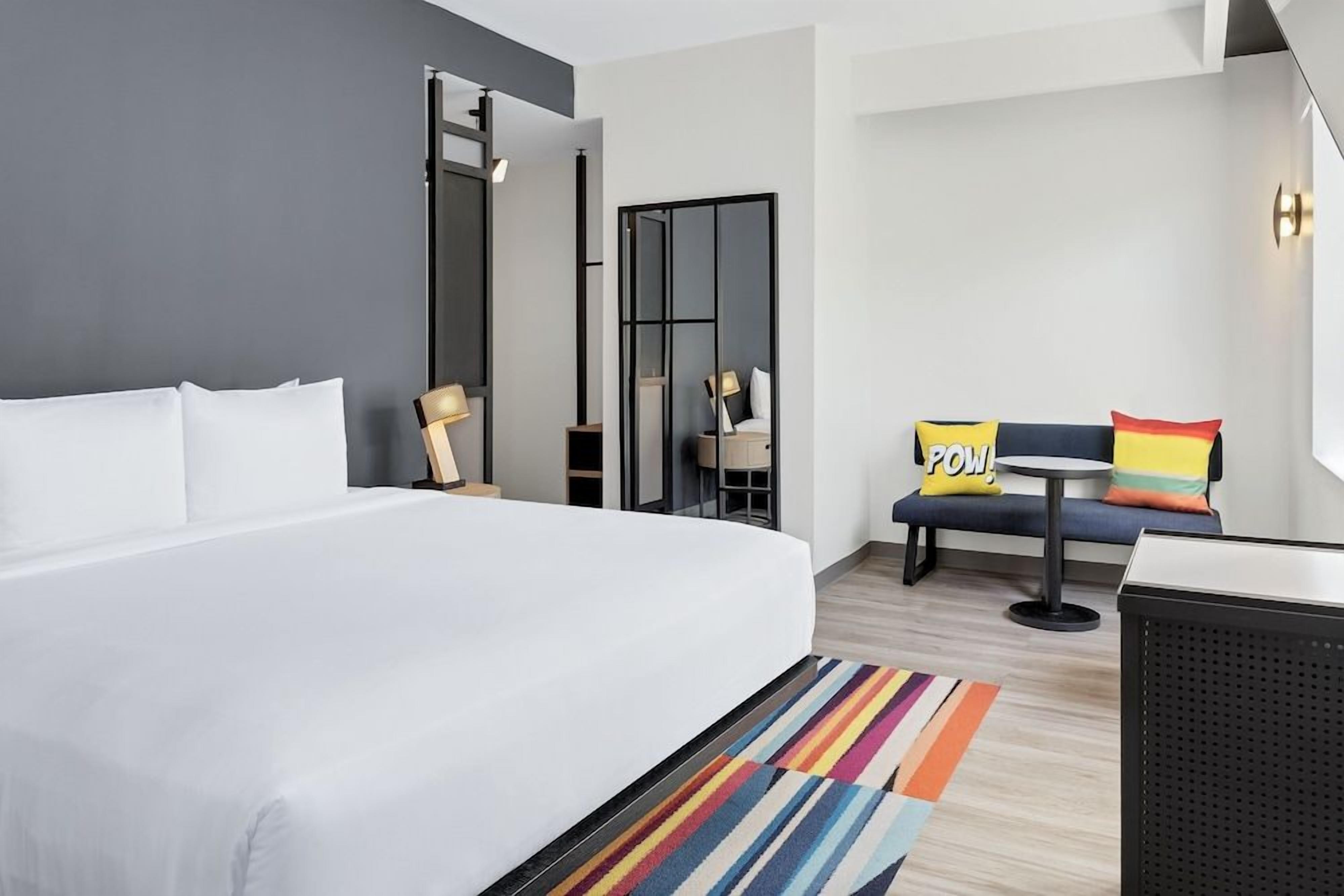 A hotel room with a large bed, a small table, a chair, and a sofa with colorful cushions.