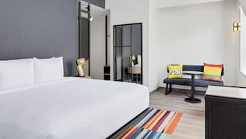 A hotel room with a large bed, a small table, a chair, and a sofa with colorful cushions.