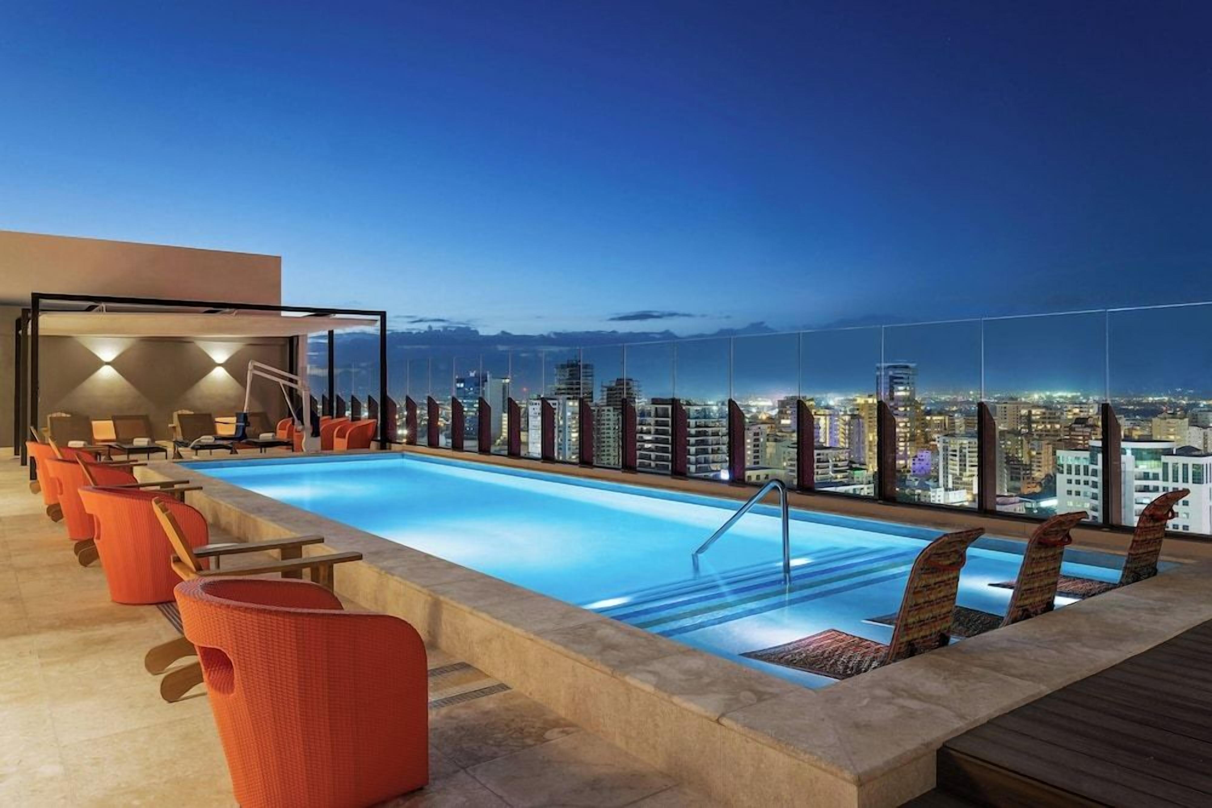 A rooftop pool with lounge chairs and a city view at dusk.