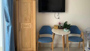 A room with a wooden wardrobe, a TV mounted on the wall, two blue chairs around a small table, and a standing fan. - Tuan Anh 2 Hotel by Zuzu (Dong Thuan)