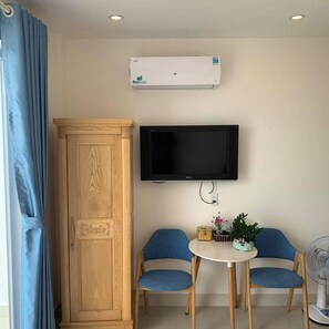 Une pièce avec une armoire en bois, un téléviseur fixé au mur, deux chaises bleues disposées autour d’une petite table et un ventilateur sur pied.