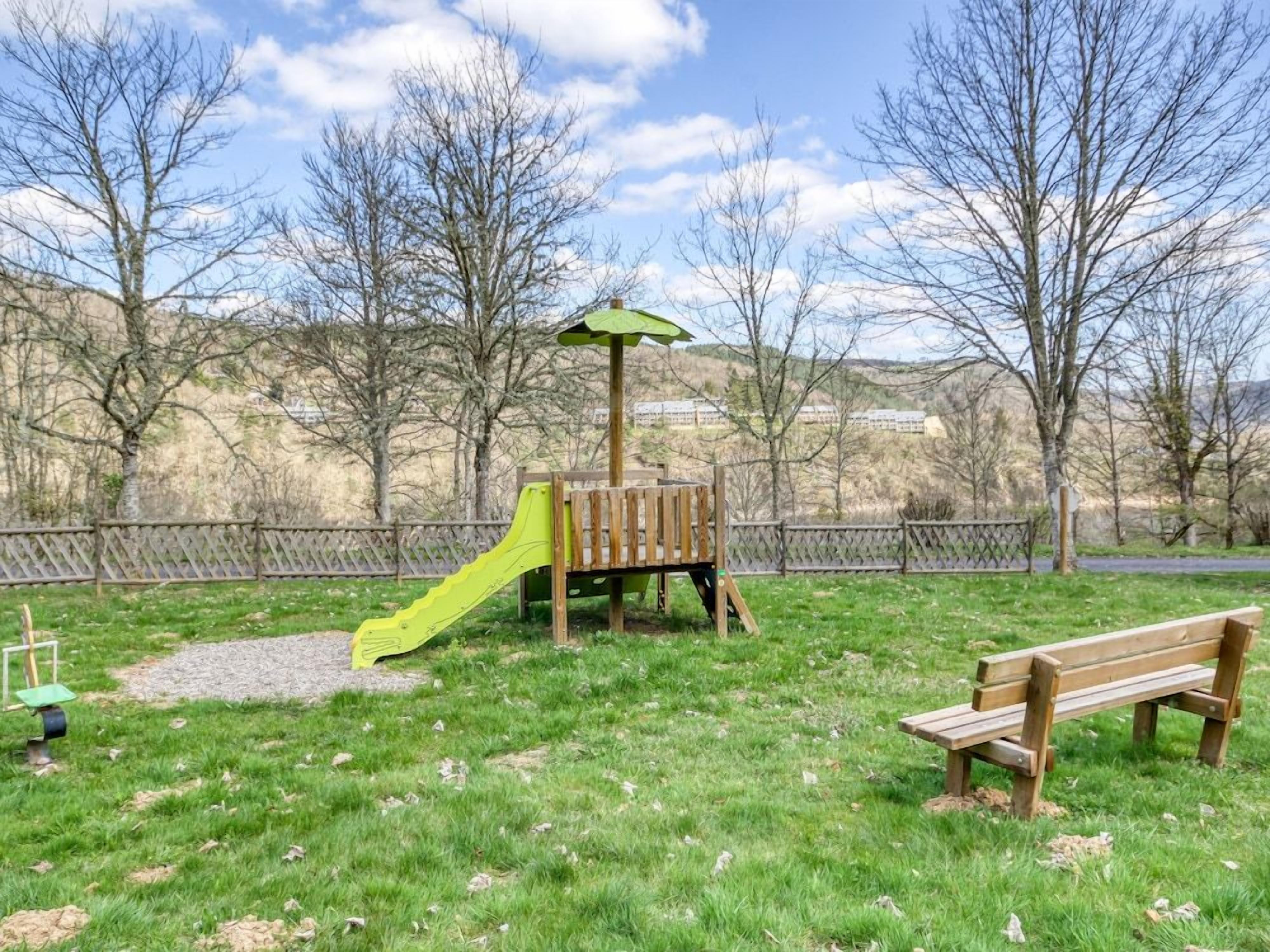 A playground with a slide and a climbing structure, surrounded by a fence and trees.