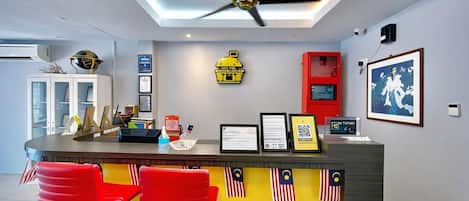 Un área de recepción con un mostrador, dos taburetes rojos, un ventilador de techo, diplomas enmarcados, un armario rojo y una cámara de seguridad instalada en la pared.