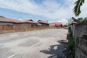A paved parking area with a brick building and a fence on the left, a red-roofed structure in the background, and a tall building in the distance. - Swing & Pillows AT Klebang, Melaka (Malacca City)