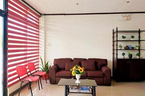 A living room with a sofa, a coffee table with a vase, red chairs, a bookshelf, and a window with red blinds. - EC Hotel Near Laoag Airport by Zuzu (Laoag)