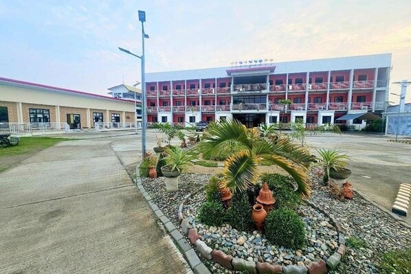 A multi-story hotel with a red facade, a parking area, and landscaped gardens with palm trees and potted plants.