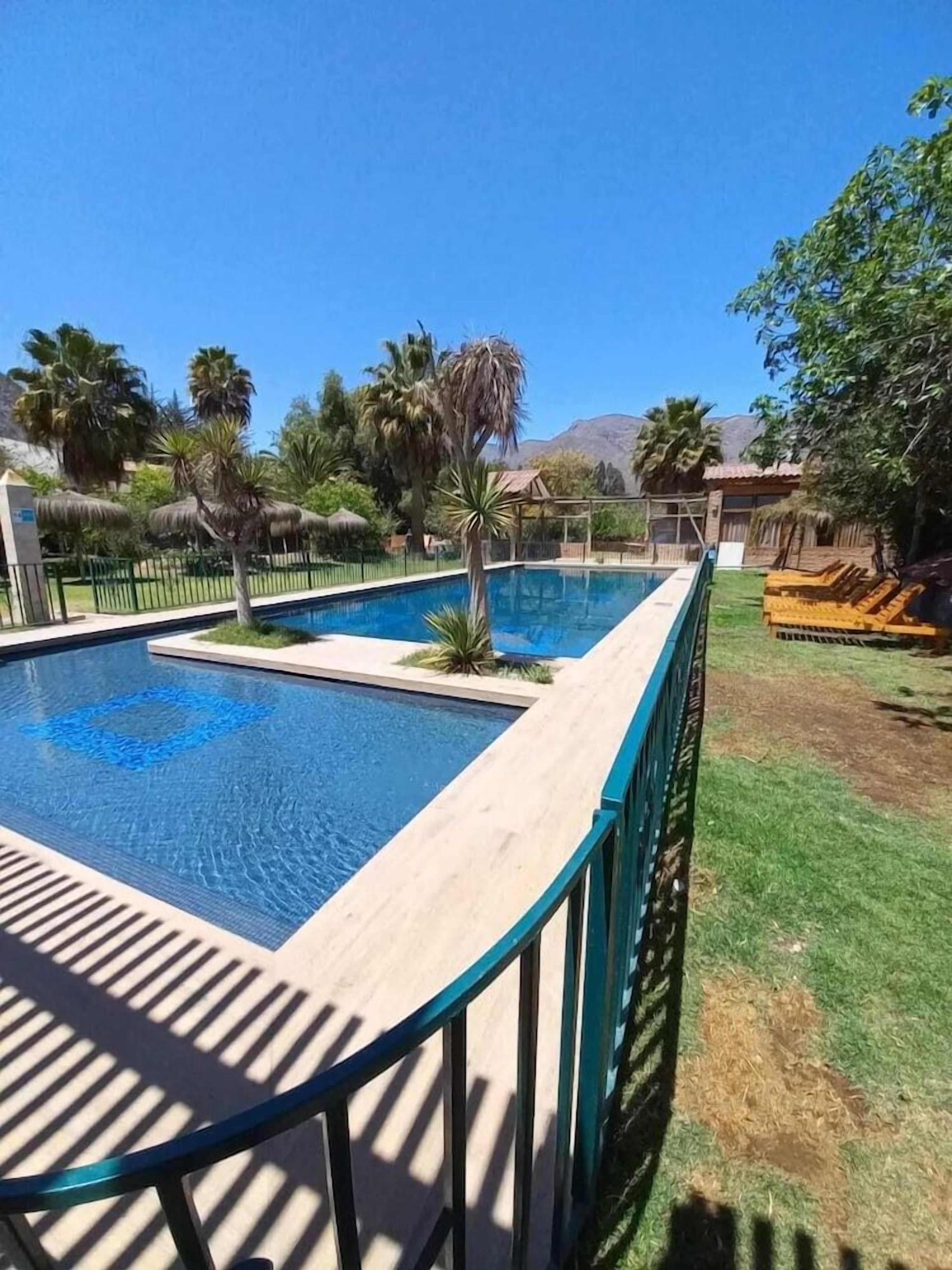 A rectangular swimming pool with a wooden deck and metal railing, surrounded by grass and palm trees.