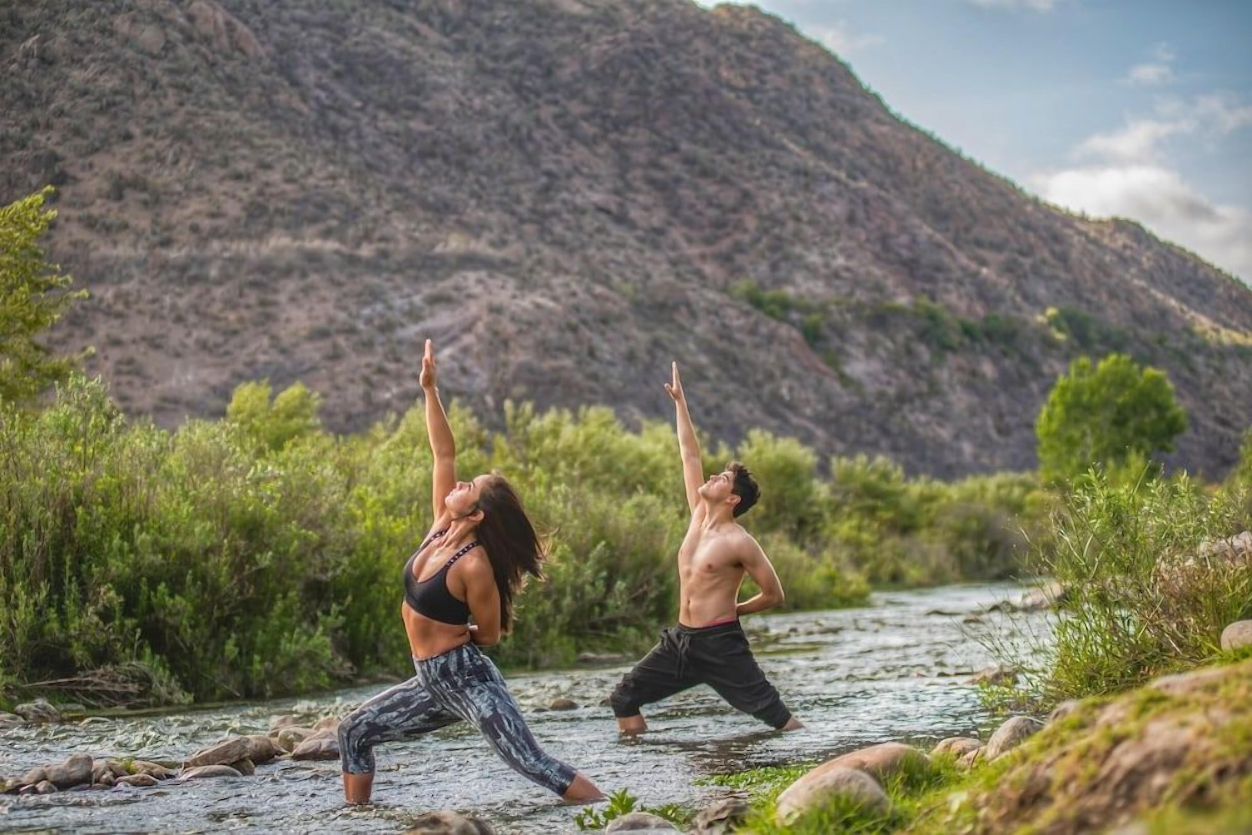 Two individuals practicing yoga on a riverbank with a mountainous backdrop.