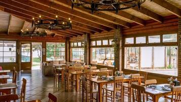 A spacious indoor dining area with wooden tables and chairs, large windows, and a high ceiling with exposed beams.