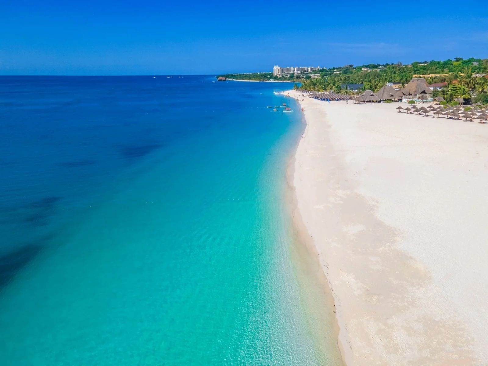 A beach with clear turquoise waters, a sandy shore, and buildings in the background.