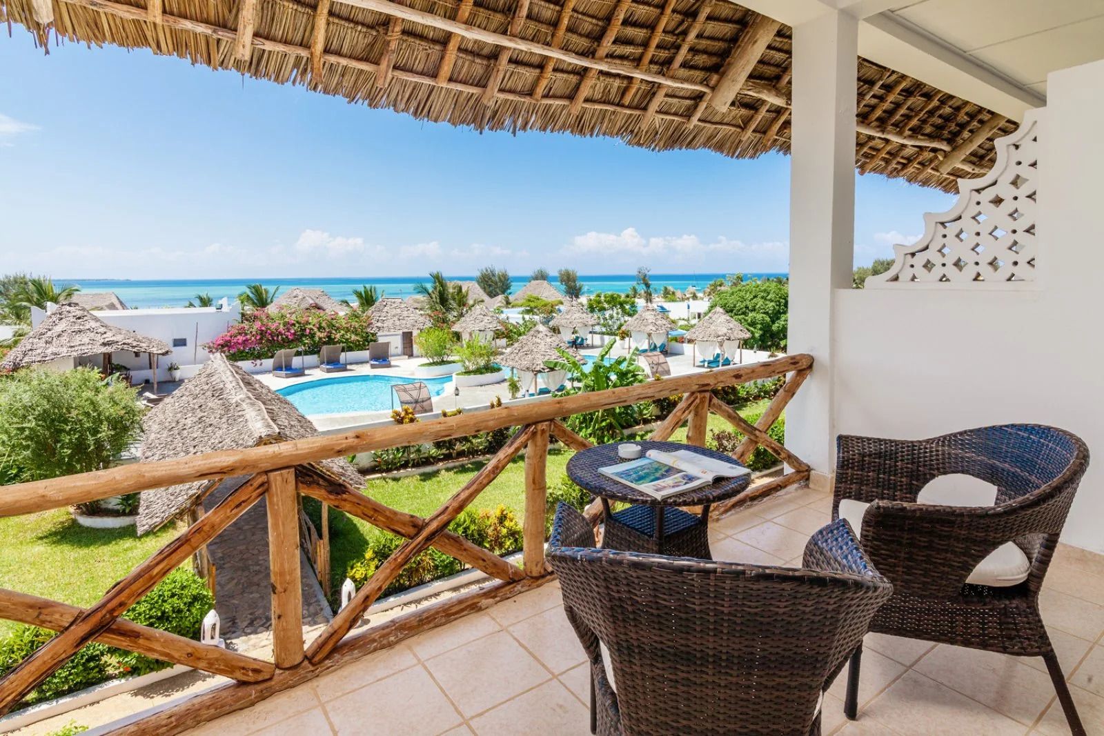 A balcony with wicker chairs, a small table, and a view of a pool and ocean.