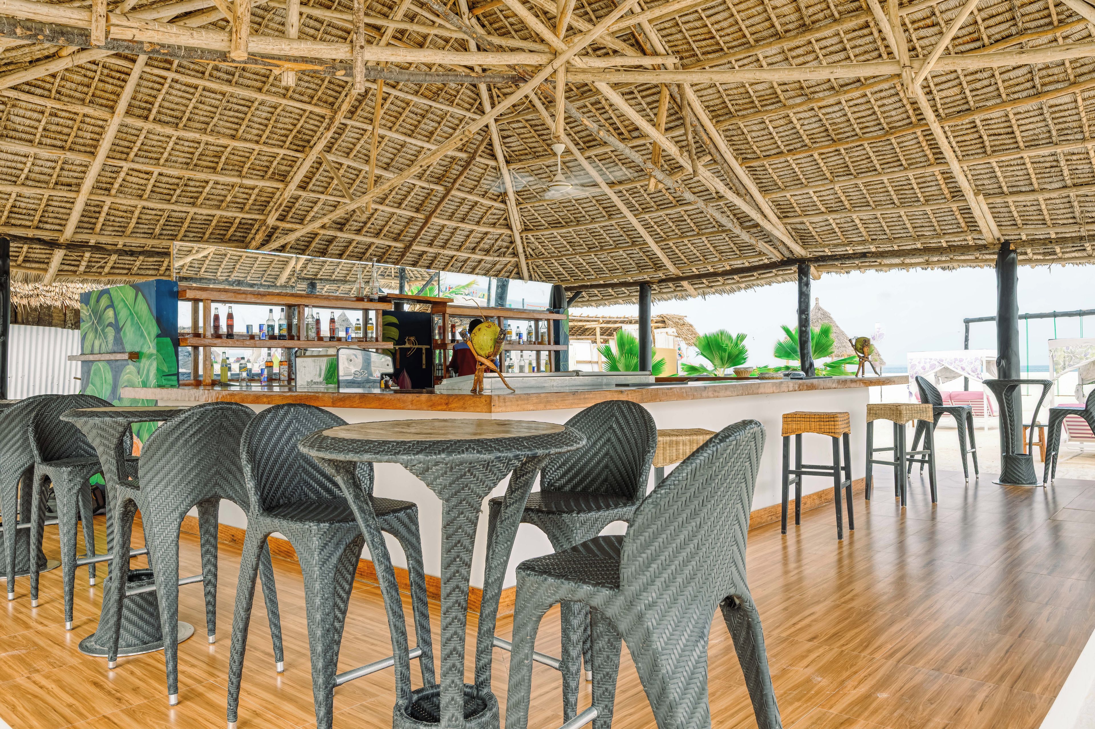 An indoor seating area with wicker chairs and tables, a bar with bottles, and a thatched roof structure.