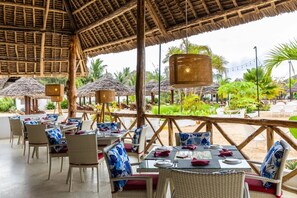 Un espace de restauration en plein air avec des chaises et des tables en osier, des parasols à toits de chaume et des lampes en osier suspendues.