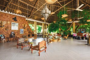 Un vaste espace intérieur doté d’un haut plafond, de poutres en bois et d’un toit de chaume. On y trouve plusieurs coins salon avec des tables et des chaises, et un grand arbre est visible à l’arrière-plan.