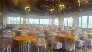 A banquet hall with round tables and chairs, large windows, and a high ceiling with hanging plants.