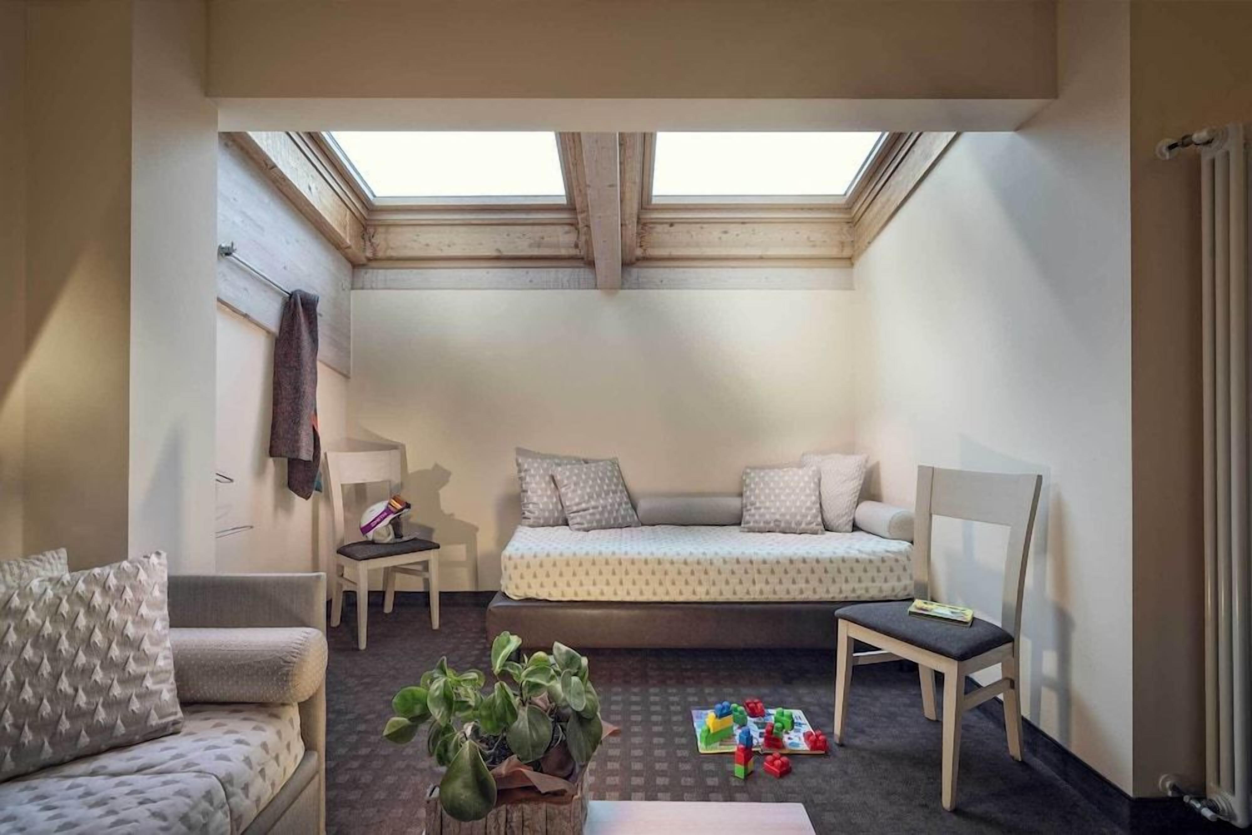 A small, cozy room with a loft bed, a desk, and a chair. There is a skylight above the bed, and a plant on the floor.