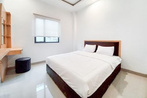 Un dormitorio con una cama, un ventanal, un estante de madera y un otoman redondo.