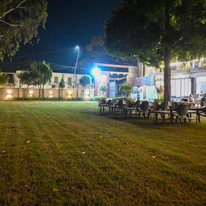 An outdoor seating area with tables and chairs, surrounded by trees and a building with illuminated windows. - Hotel Kedar Dham (Mathura)