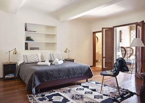 A bedroom with a bed, bedside tables, a chair, a bookshelf, and a door leading to another room.
